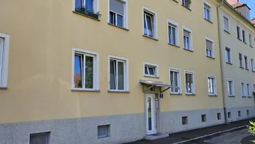 Mehrstöckiges Wohngebäude mit gelber Fassade, weißen Fenstern und Rollläden an einer ruhigen Straße.