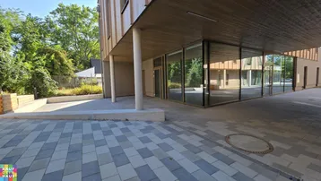 Moderner Gebäudeeingang mit großen Glasfronten und gepflastertem Vorplatz, umgeben von Bäumen und Grünflächen.