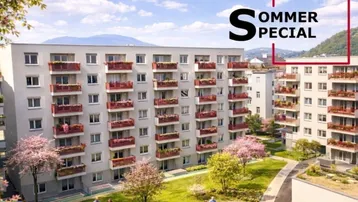 Modernes Mehrfamilienhaus mit roten Balkonen und gepflegter Grünanlage, umgeben von Bergen.