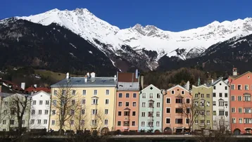 Bunte Häuserfassaden mit traditioneller Architektur vor einer beeindruckenden Kulisse schneebedeckter Berge unter blauem Himmel.