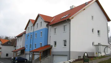 Modernes Mehrfamilienhaus mit blauer Fassadenakzentuierung, Garagen und Satteldach in ländlicher Umgebung.