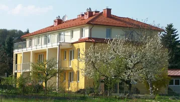 Mehrfamilienhaus mit gelber Fassade, roten Ziegeldächern und Balkonen in ländlicher Umgebung.