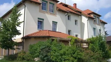 Gepflegtes Mehrfamilienhaus mit weißer Fassade, rotem Satteldach und grünen Rollläden, umgeben von Bäumen und Hecken.