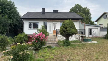 Einfamilienhaus mit weißer Fassade, schwarzem Dach und einem ungepflegten Vorgarten mit Rosenbüschen.