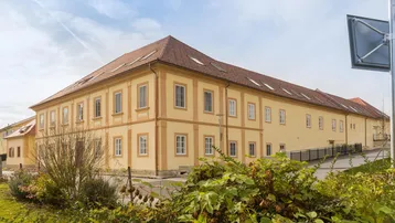 Gelbes Gebäude mit traditionellem Ziegeldach und vielen Fenstern, umgeben von gepflegten Grünflächen unter blauem Himmel.