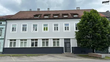 Graue Fassade eines Mehrfamilienhauses mit vielen Fenstern und einem Baum davor.