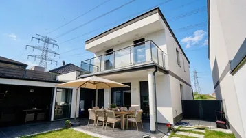 Moderne Hausfassade mit großer Terrasse, Gartenbereich und Sitzgelegenheiten unter einem Sonnenschirm.