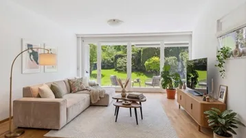 Helles Wohnzimmer mit beigem Sofa, Holzboden und großen Fenstern mit Blick in den Garten.