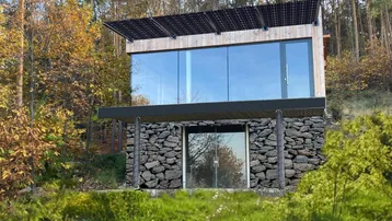 Modernes Haus mit großer Glasfassade, Steinsockel und Solarpaneelen auf dem Dach, umgeben von Bäumen.