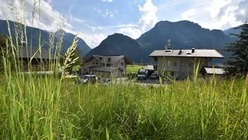 Ländliche Umgebung mit Häusern, umgeben von grünen Wiesen und Bergen unter blauem Himmel.