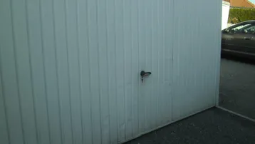 Weiße Garagentür mit vertikalen Rillen und einem Griff, die leichte Dellen aufweist. Ein Auto ist im Hintergrund sichtbar.