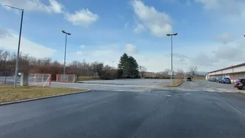 Großer asphaltierter Parkplatz oder Industriegelände mit angrenzendem Gebäude und einigen Fahrzeugen unter bewölktem Himmel.
