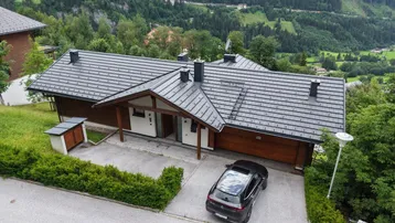 Luftaufnahme eines modernen Hauses mit dunklem Dach und gepflegter Auffahrt in bergiger Umgebung.