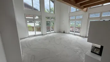 Geräumiger Wohnbereich im Rohbau mit hohen Decken, großen Fenstern und modernem Kamin.