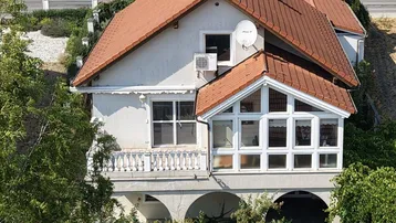 Ansicht eines Hauses mit rotem Ziegeldach, weißer Fassade, Balkon und einem verglasten Wintergarten.