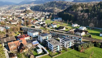 Luftaufnahme einer Siedlung mit modernen Gebäuden, grünen Wiesen und bewaldeten Hügeln im Hintergrund.