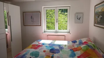 Helles Schlafzimmer mit farbenfroher Bettwäsche und einem Fenster, das den Blick ins Grüne freigibt.