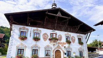 Traditionelles Gebäude mit kunstvollen Fassadenmalereien, Holzbalkonen und Blumenkästen unter einem blauen Himmel.