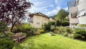 Weitläufiger Garten mit Rasen, Sitzbank und Blick auf die umliegenden Gebäude und Berge.
