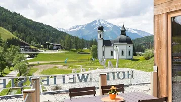Geräumige Holzterrasse mit modernen Esstischen und Stühlen, bietet einen malerischen Blick auf schneebedeckte Berge und eine Kirche.