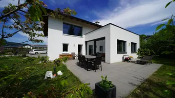 Moderne Hausfassade mit großer gepflasterter Terrasse, Garten und Essbereich im Freien unter blauem Himmel.