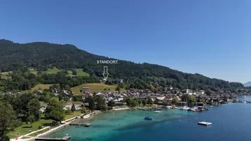 Luftaufnahme eines malerischen Ortes am Seeufer mit bewaldeten Bergen im Hintergrund und markiertem Standort.