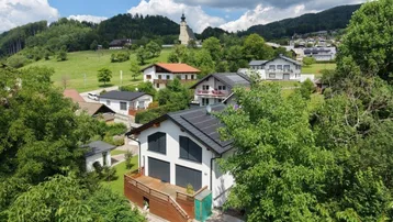 Luftaufnahme eines modernen Hauses in einer grünen Hügellandschaft mit Blick auf ein Dorf und eine Kirche.