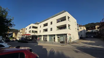 Modernes weißes Gebäude mit Balkonen und Geschäftsflächen im Erdgeschoss an einer Straße gelegen.