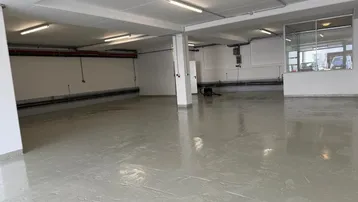 Geräumige, leere Gewerbefläche mit hellem Industrieboden, weißen Wänden und sichtbaren Installationen an der Decke.