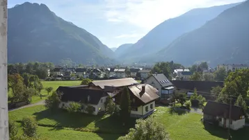 Weitläufiger Blick über grüne Felder und ein Dorf mit Bergen im Hintergrund unter blauem Himmel.