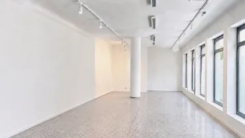 Heller, leerer Geschäftsraum mit großen Fenstern, Terrazzoboden und moderner Schienenbeleuchtung an der Decke.