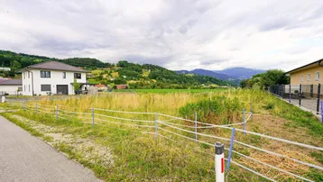 Blick auf ein unbebautes Grundstück mit umliegenden Häusern und einer grünen Hügellandschaft im Hintergrund.