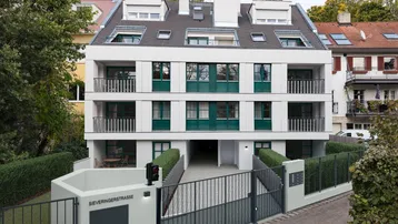 Modernes Mehrfamilienhaus mit weißer Fassade, grünen Fensterläden und Balkonen, umgeben von gepflegten Hecken.
