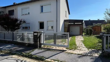 Weißes Einfamilienhaus mit Vorgarten, Zaun und Einfahrt unter blauem Himmel.