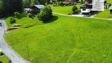 Luftaufnahme eines grünen Baugrundstücks in einer bergigen Wohngegend mit umliegenden Häusern und viel Natur.