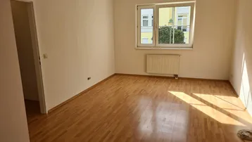 Helles Zimmer mit Parkettboden und Fenster, das viel Tageslicht hereinlässt.