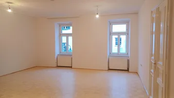 Helles, leeres Wohnzimmer mit Parkettboden und zwei Fenstern, die viel Tageslicht hereinlassen.