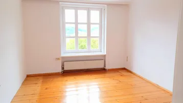 Helles Zimmer mit Holzdielenboden und großem Fenster, das viel Tageslicht hereinlässt.