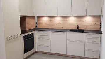 Moderne L-förmige Einbauküche mit hellen Fronten, Holzrückwand und integriertem Backofen.