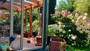 Heller Wintergarten mit vielen Pflanzen, Blick in den blühenden Garten und Zugang zur Terrasse.