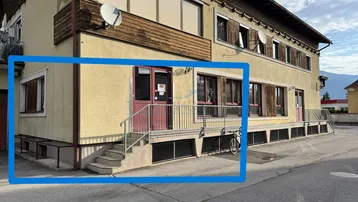 Gebäudeaußenansicht mit mehreren Fenstern, einem Eingangsbereich und einer kleinen Treppe mit Geländer.