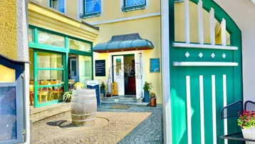Eingangsbereich eines charmanten Gebäudes mit gelber Fassade und grünem Tor, gepflastertem Innenhof und Holzbottich.