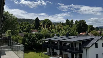 Blick von einem modernen Balkon mit Glasgeländer auf eine grüne Umgebung und benachbarte Gebäude.