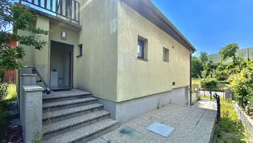 Eingangsbereich des Hauses mit Außentreppe und umliegender Vegetation unter strahlendem Himmel.