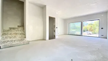 Geräumiger, unfertiger Innenraum mit großen Fenstern und Treppenaufgang, bereit für den Ausbau.