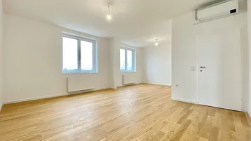 Heller, leerer Wohnraum mit Holzboden, weißen Wänden und großen Fenstern, die viel Tageslicht hereinlassen.