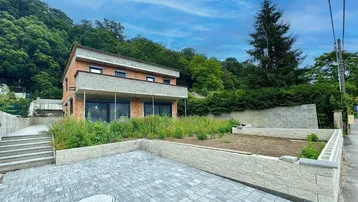 Neubauhaus in Hanglage mit gepflastertem Vorplatz und vorbereiteten Gartenbeeten, umgeben von viel Grün.