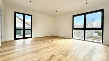Geräumiges, leeres Zimmer mit hellem Holzboden und großen Fenstern, die viel Tageslicht hereinlassen.