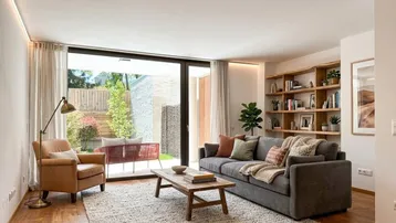 Helles Wohnzimmer mit großen Fenstern, Holzboden und modernem Sofa, das Zugang zu einem Außenbereich bietet.