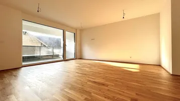 Helles, leeres Wohnzimmer mit großen Fenstern, Holzboden und Zugang zum Balkon.